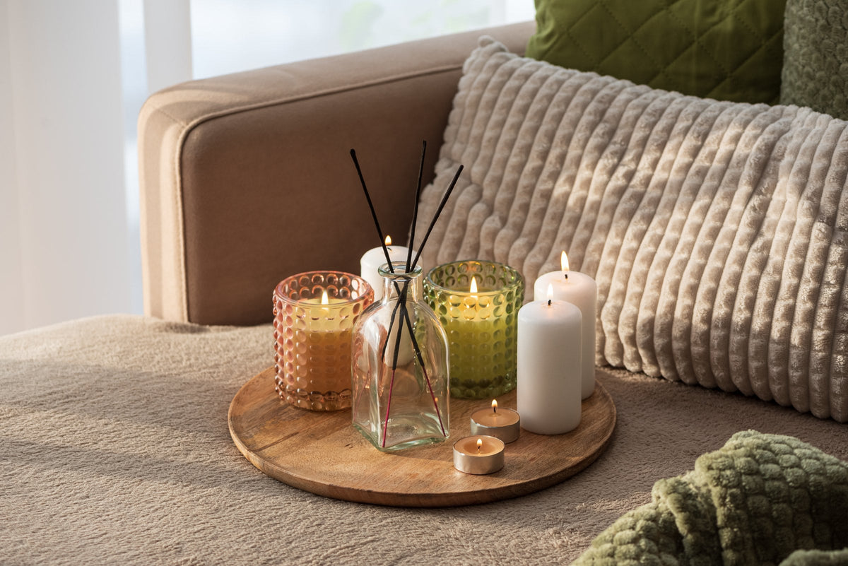 Candles and a reed diffuser on a wooden tray among cushions