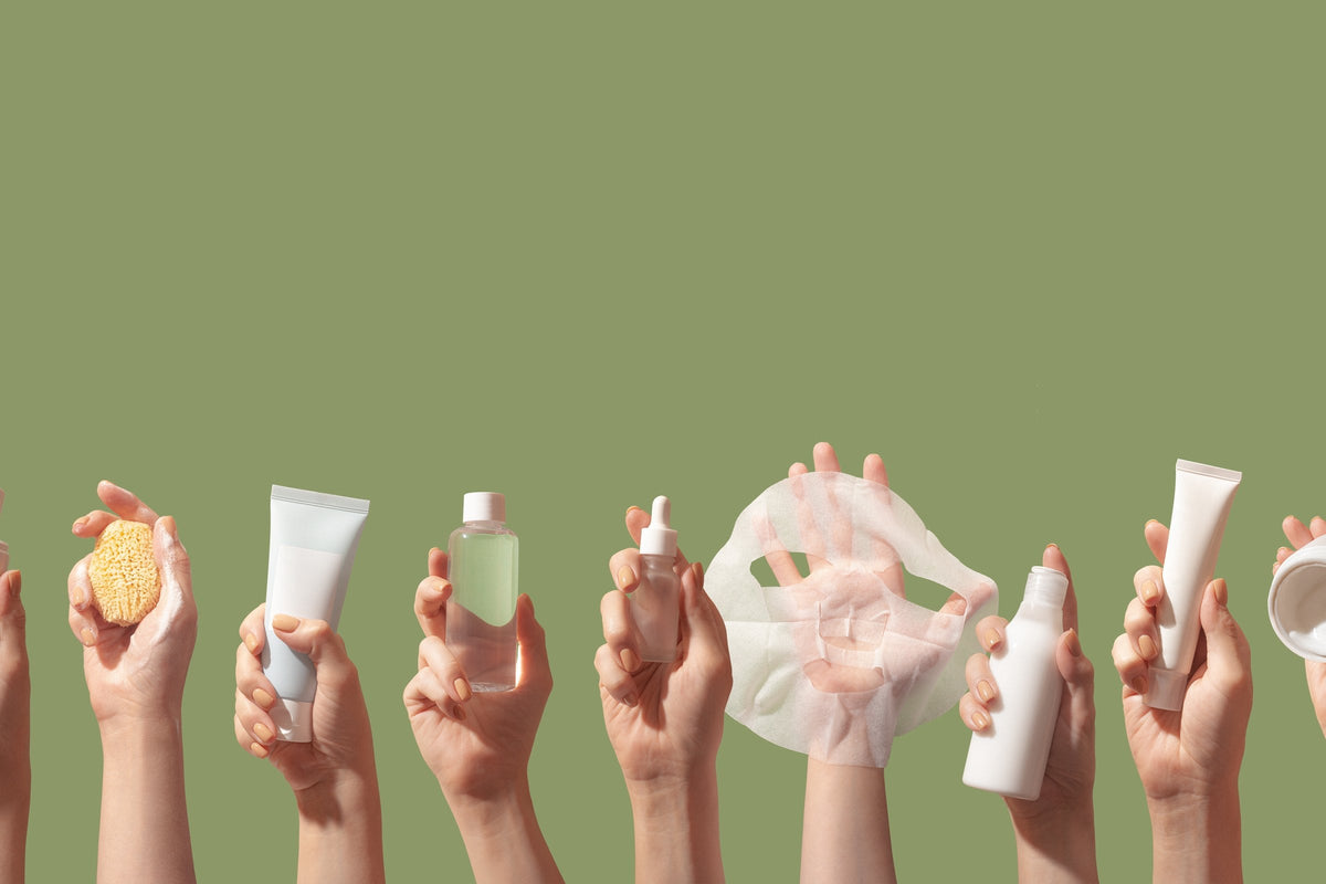 A row of hands each holding a skincare product, from cleanser and toner to a sheet mask and cream, against a green background